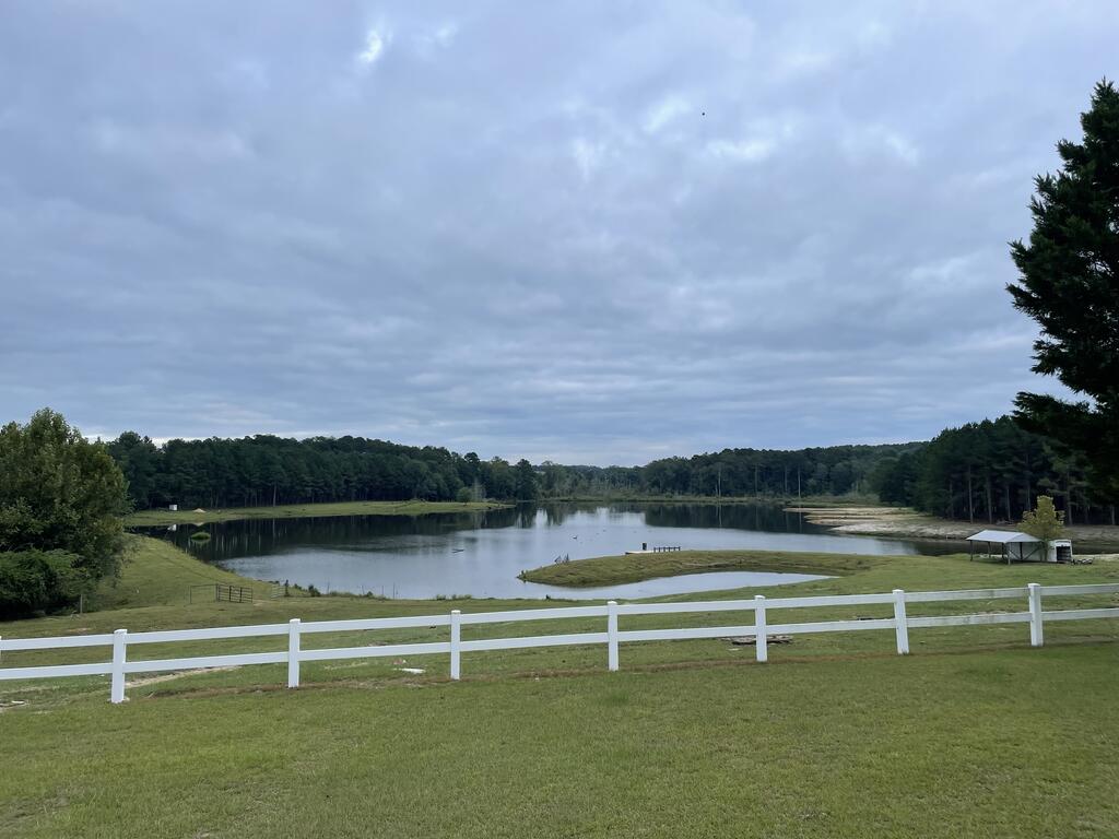Pond and wooded shoreline beyond a white fence and grassy lawn.