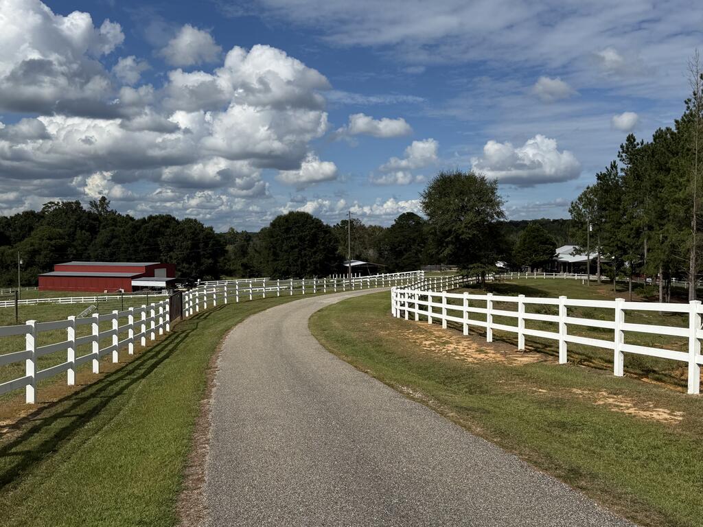 A fenced gravel drive curves toward a rural property, with a red barn and trees.