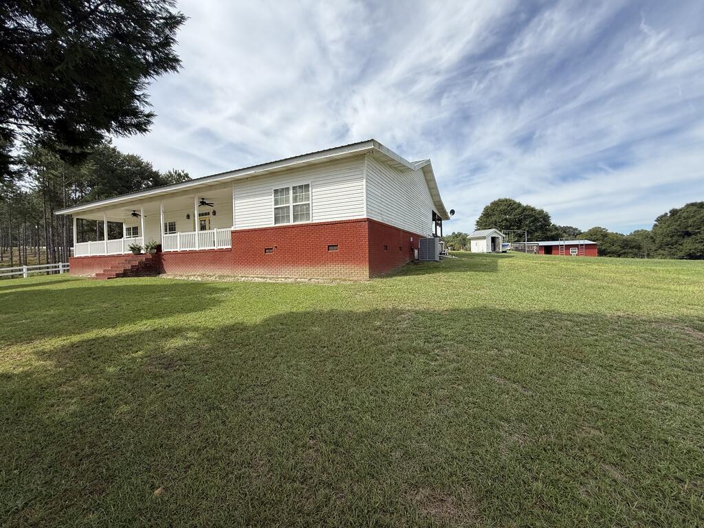 White-sided home with a red brick foundation and covered porch beside a broad lawn and outbuildings.