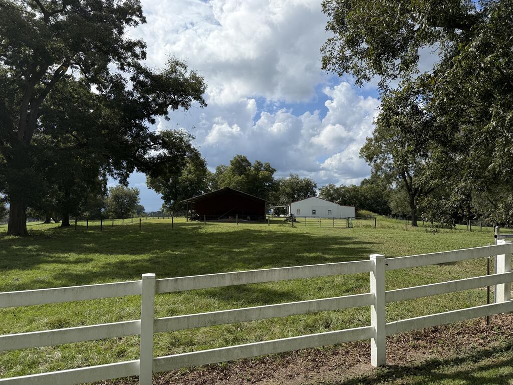Rural property with a white rail fence, grassy grounds, mature trees, and two outbuildings.