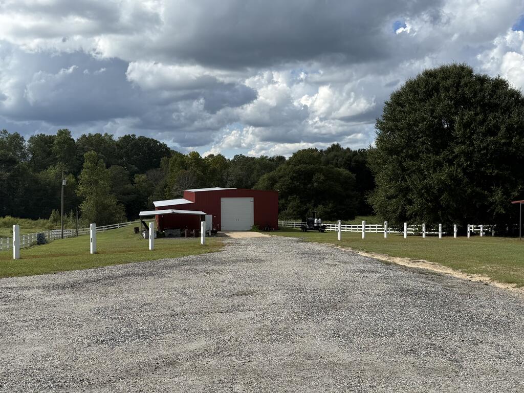 Red metal barn with a gravel driveway, white fencing, and mature trees.