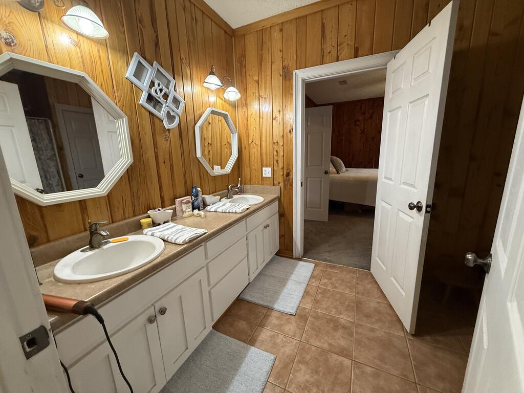 Bathroom with a double-sink vanity, wood-paneled walls, and an open doorway to a bedroom.