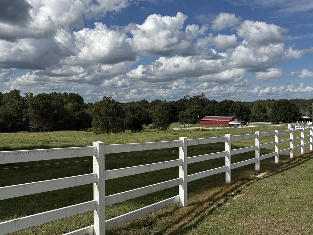 Rural pasture with white rail fencing and a red barn beneath a cloud-filled sky.