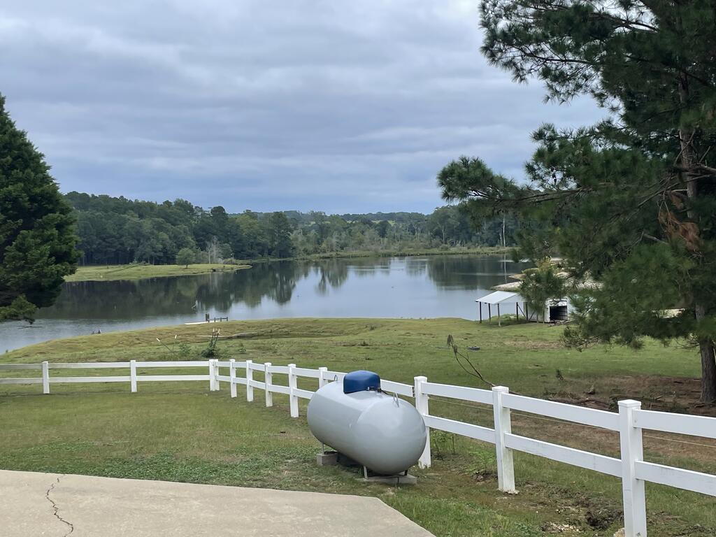 Grassy property overlooking a lake, with a white fence and propane tank in the foreground.