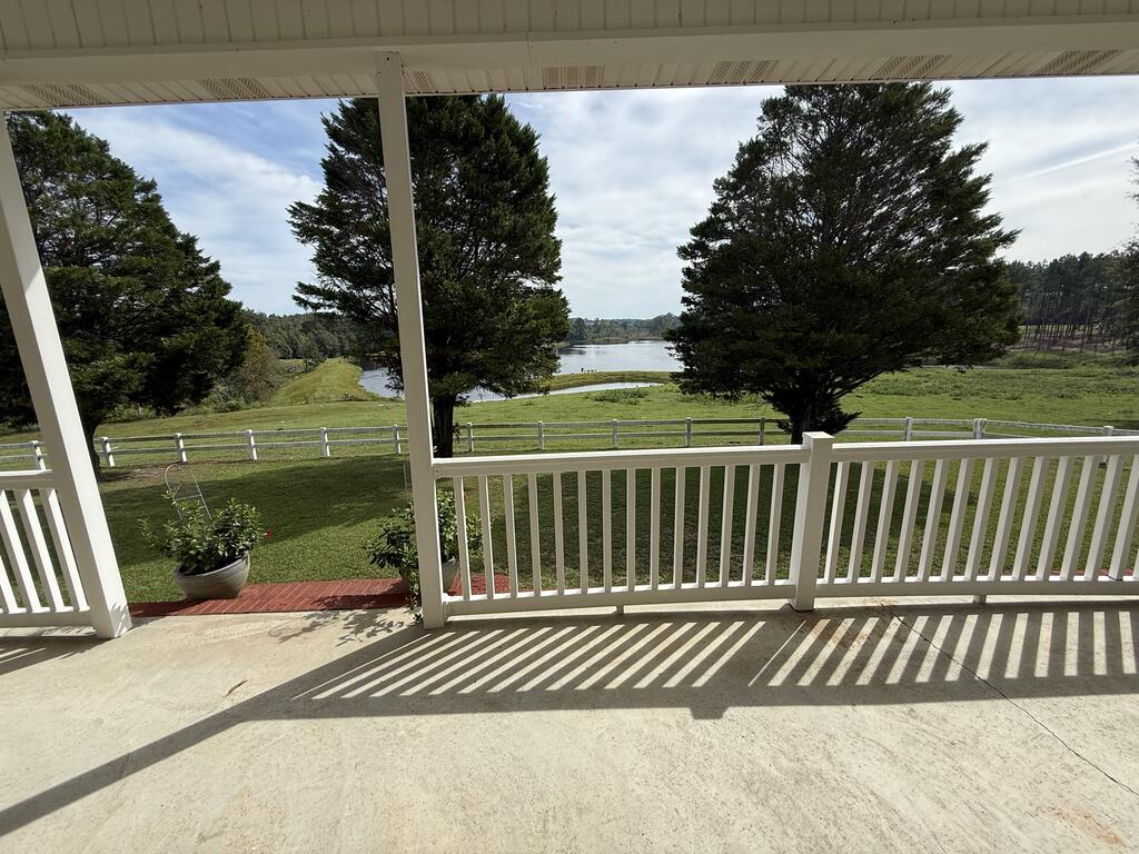 Covered porch overlooking a lake, with white railings, lawn, and evergreen trees.