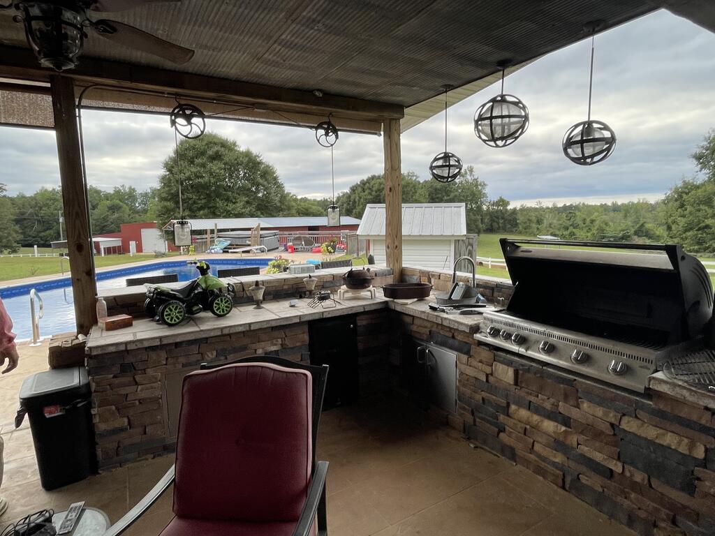 Covered outdoor kitchen with a stone counter and built-in grill overlooking a swimming pool.