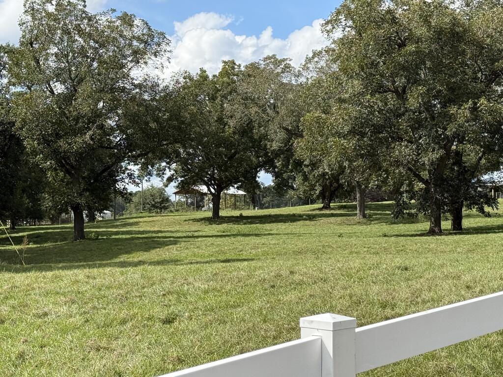 Grassy property grounds with mature trees, a white fence, and a covered shelter in the distance.