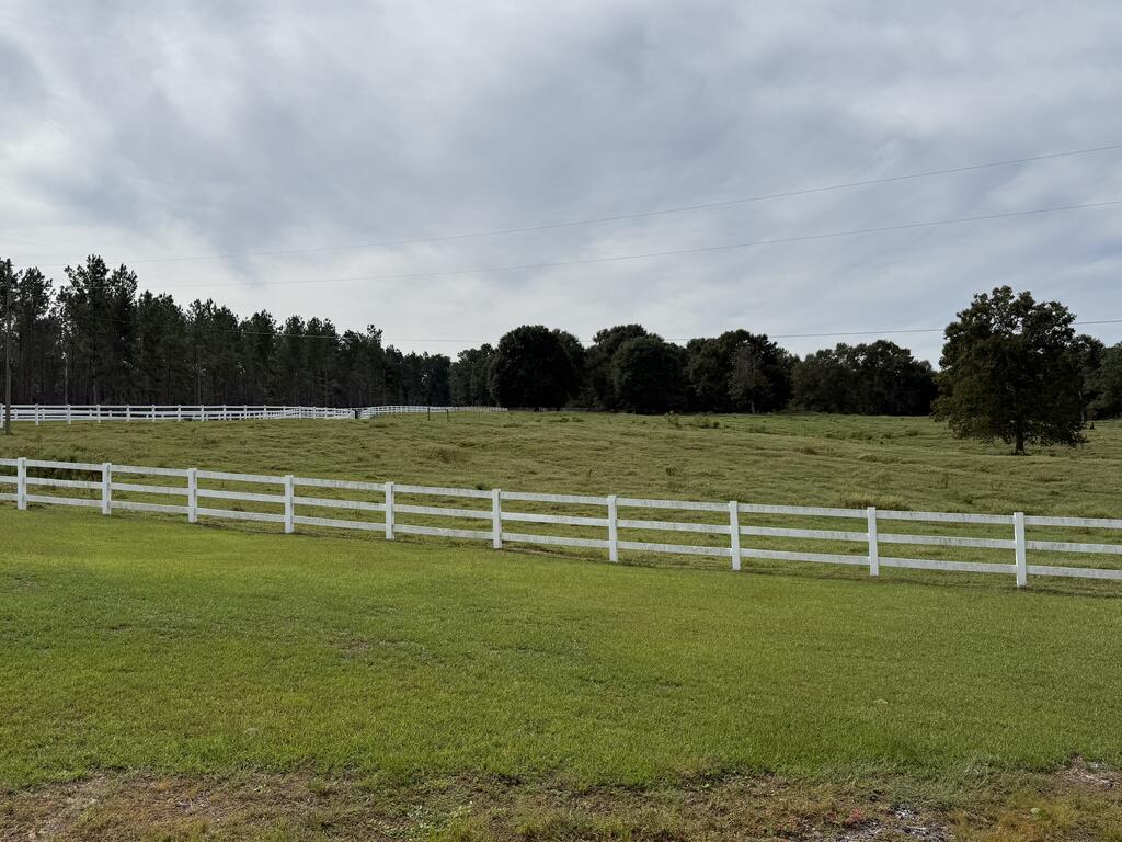 Open grassy pasture with white fencing and mature trees beneath a cloudy sky.