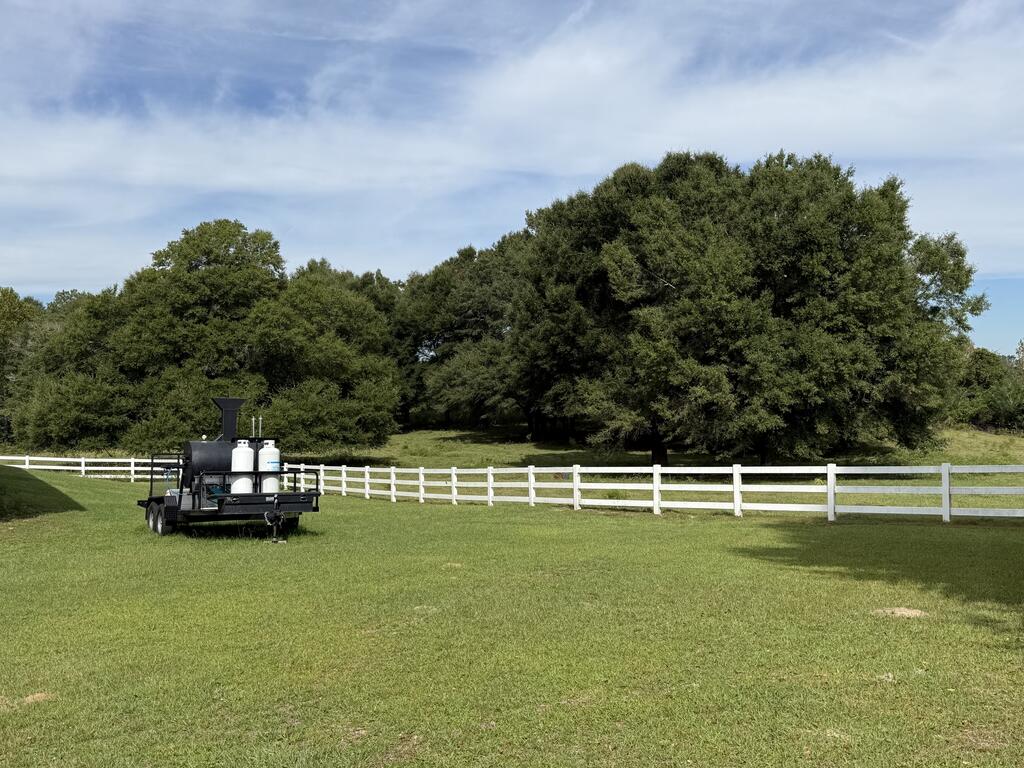 Grassy rural yard with a white rail fence, mature trees, and an equipment trailer.