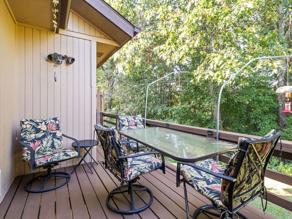 Wooden deck with a glass-top dining table and floral chairs overlooking leafy trees.