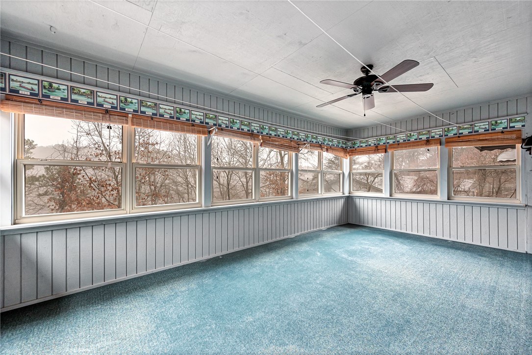 Empty sunroom with blue carpet, wraparound windows, and a ceiling fan.