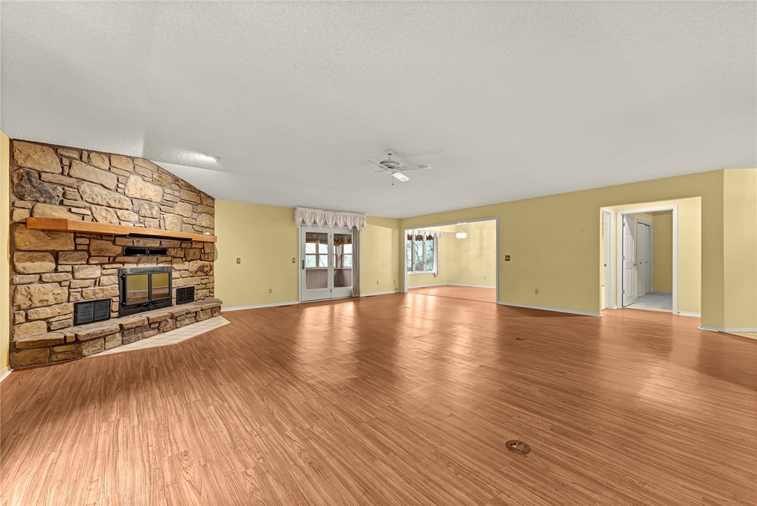 Vacant living room with a large stone fireplace, wood-look floors, and an open adjoining room.