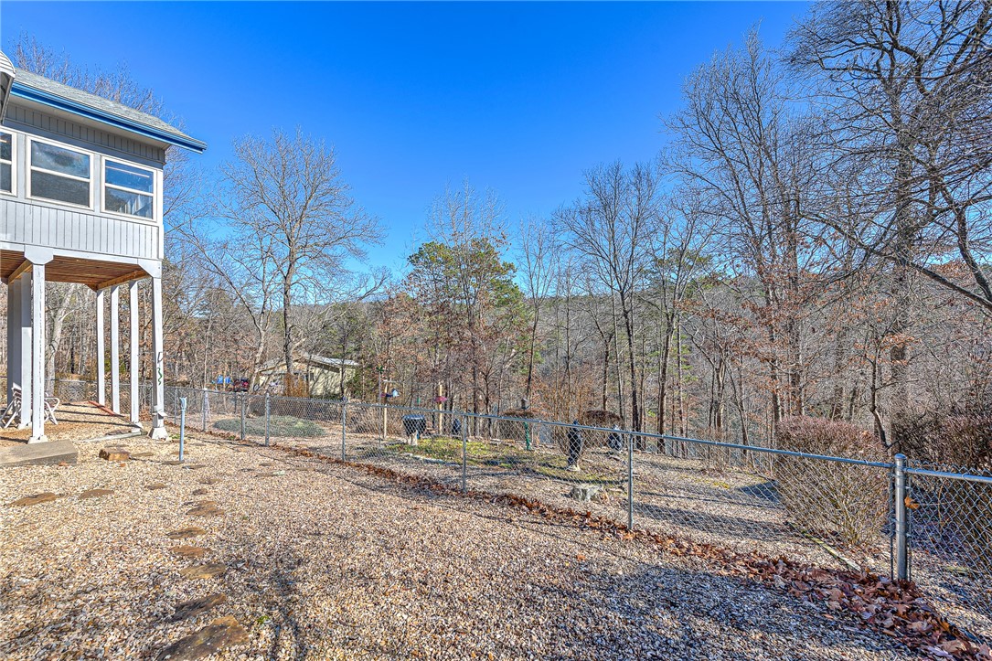 Gravel backyard with chain-link fencing and a wooded hillside behind an elevated gray house.