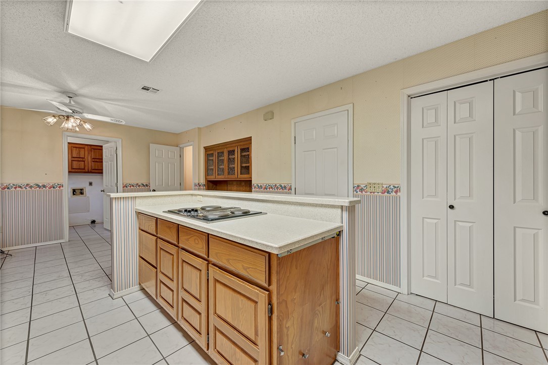 Kitchen with a wood-cabinet island and cooktop, white counters, and light tile flooring.