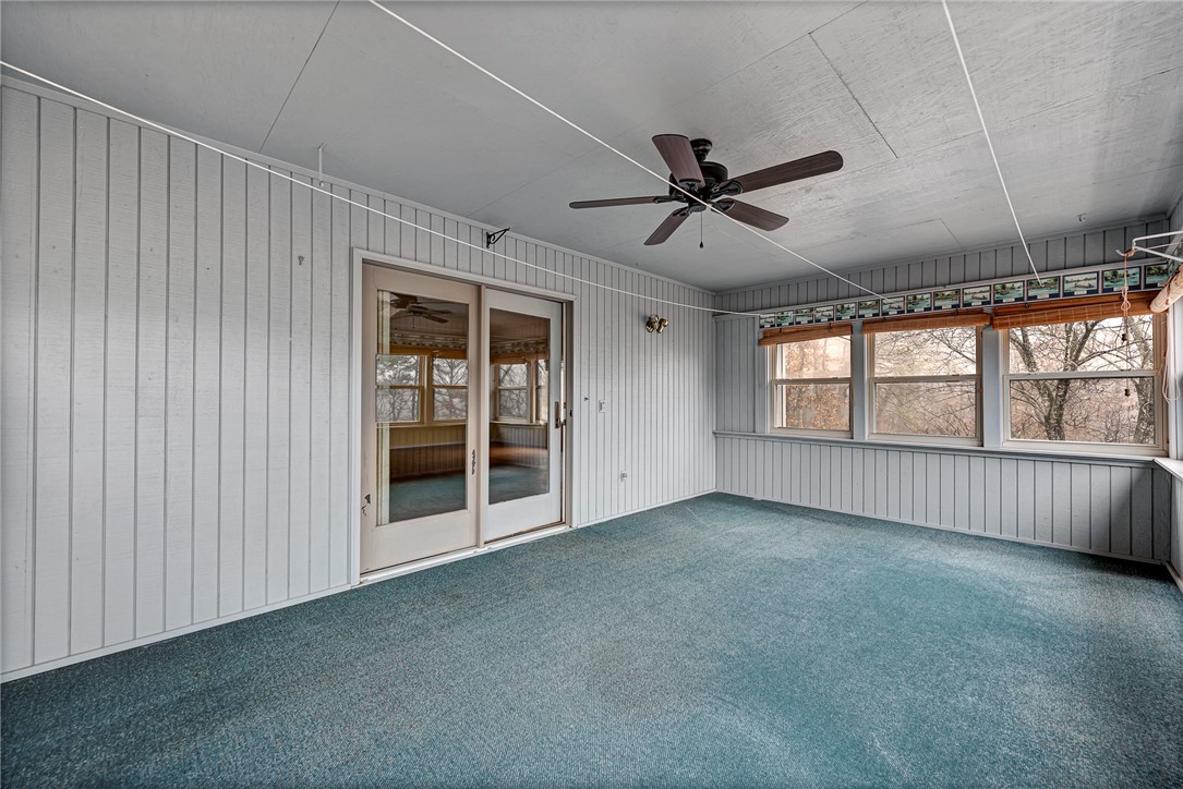 Empty sunroom with blue-green carpet, paneled walls, ceiling fan, and windows overlooking leafless trees.