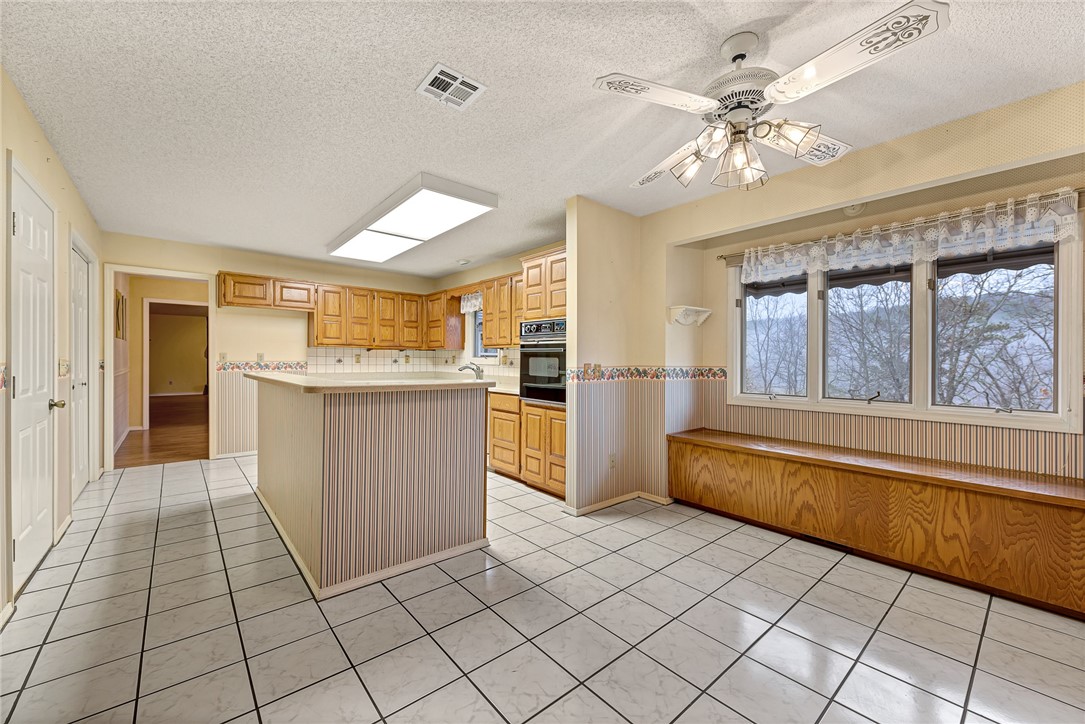 Kitchen with honey-colored cabinets, striped peninsula, tiled floor, and a built-in bench beneath wide windows.