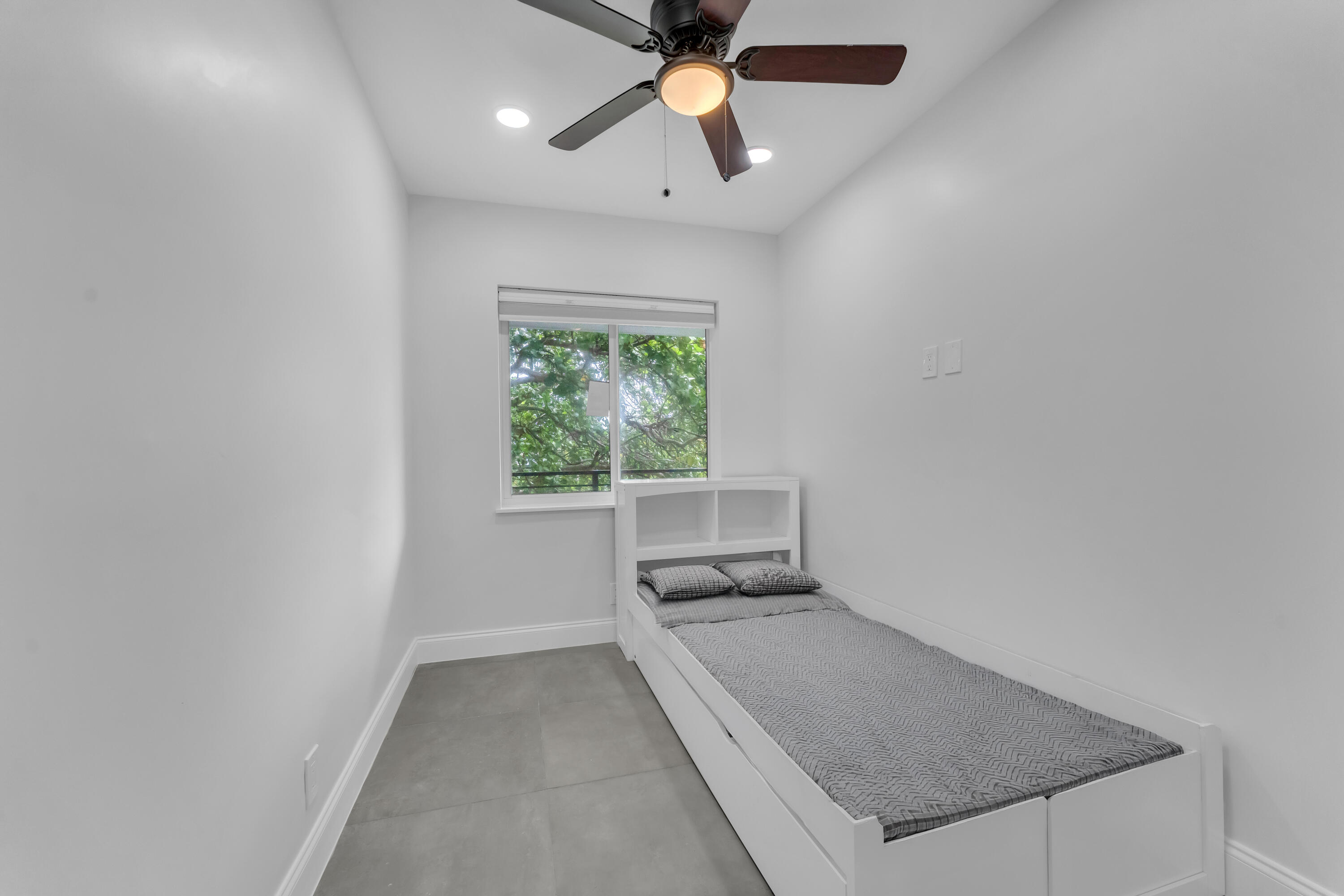 Narrow bedroom with a white single bed, gray tile floor, ceiling fan, and window overlooking leafy trees.