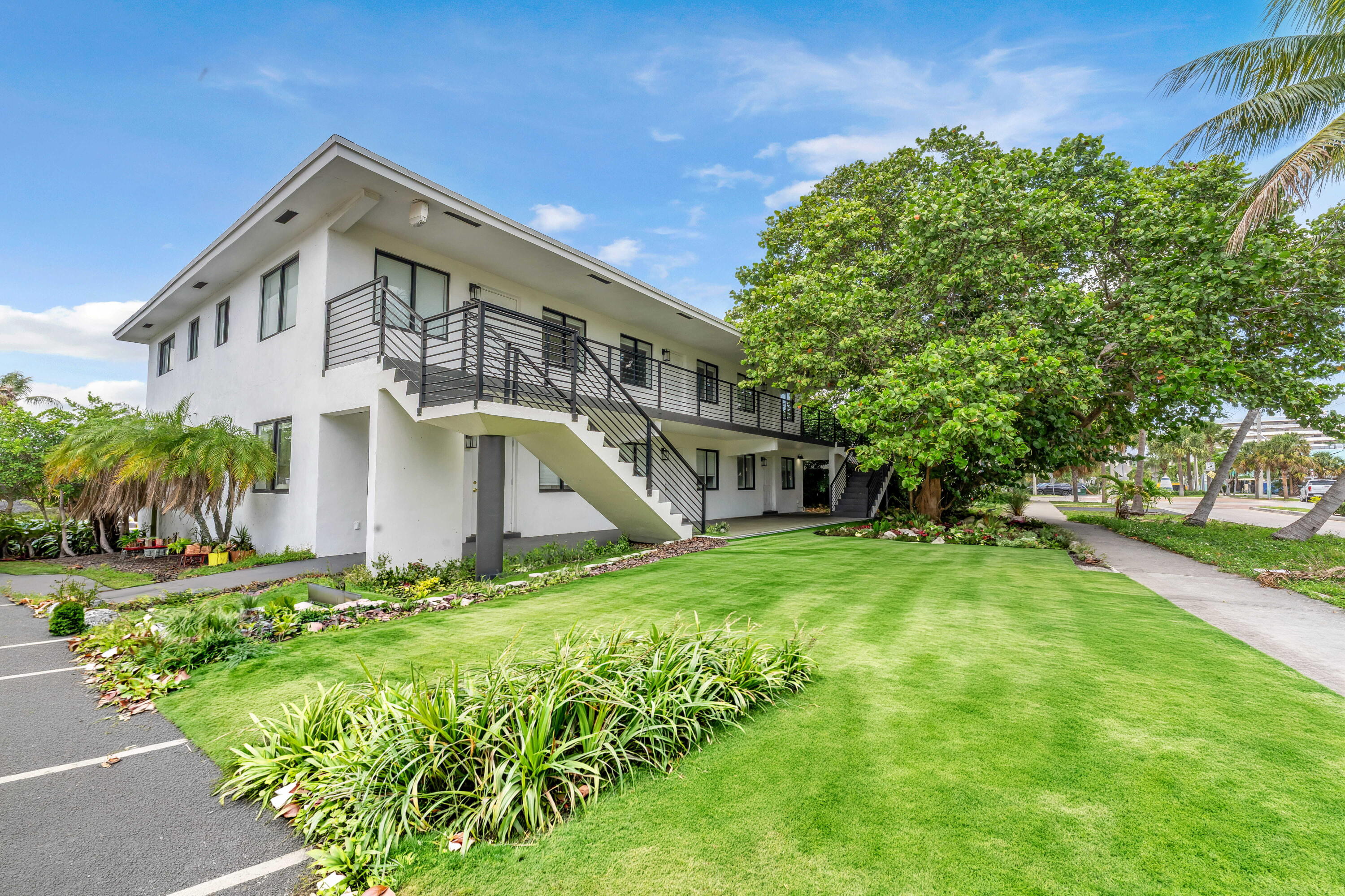 Two-story white residence with a long balcony, exterior staircase, landscaped lawn, and tropical trees.