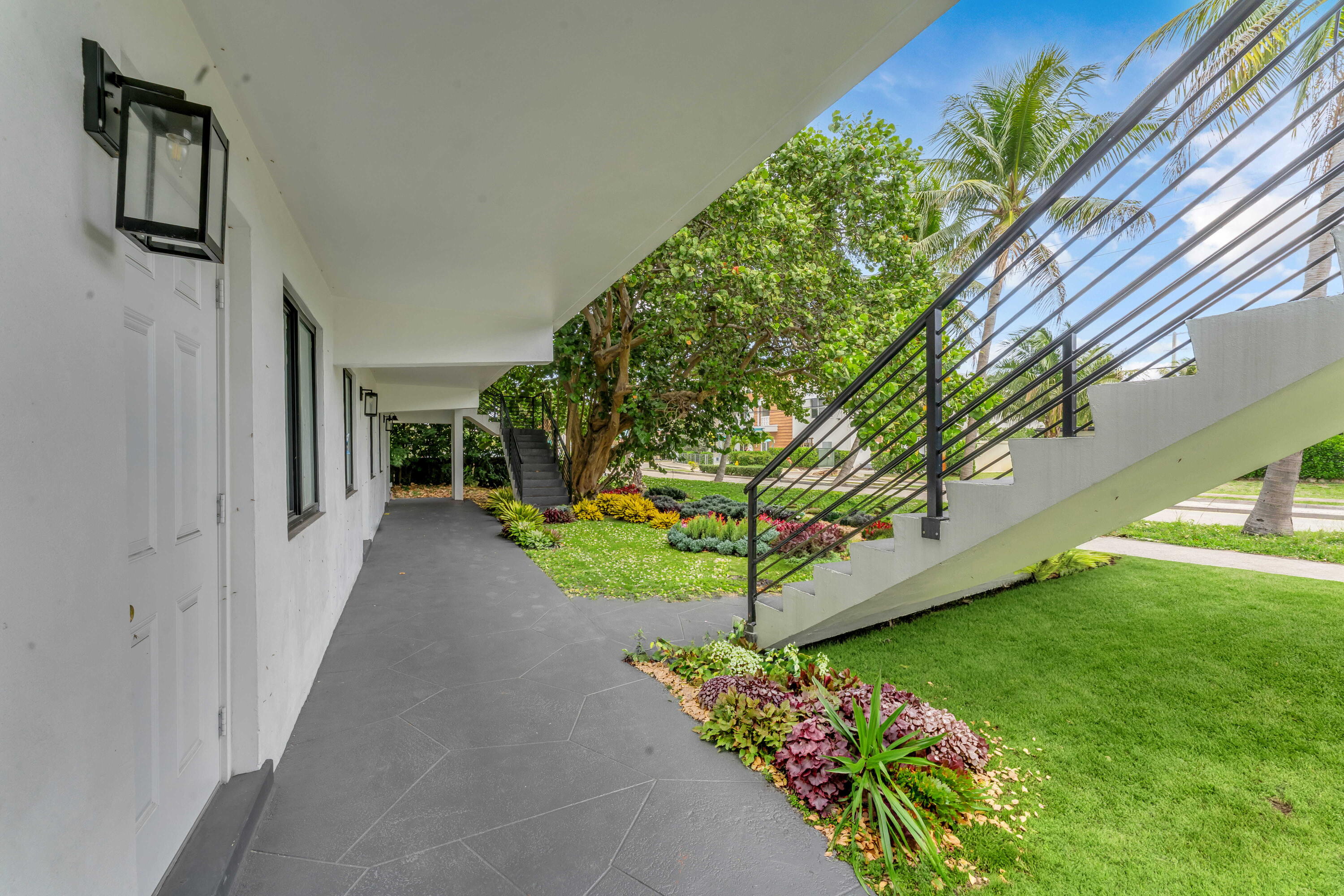 White building exterior with a covered walkway, black metal stairs, and tropical landscaping.