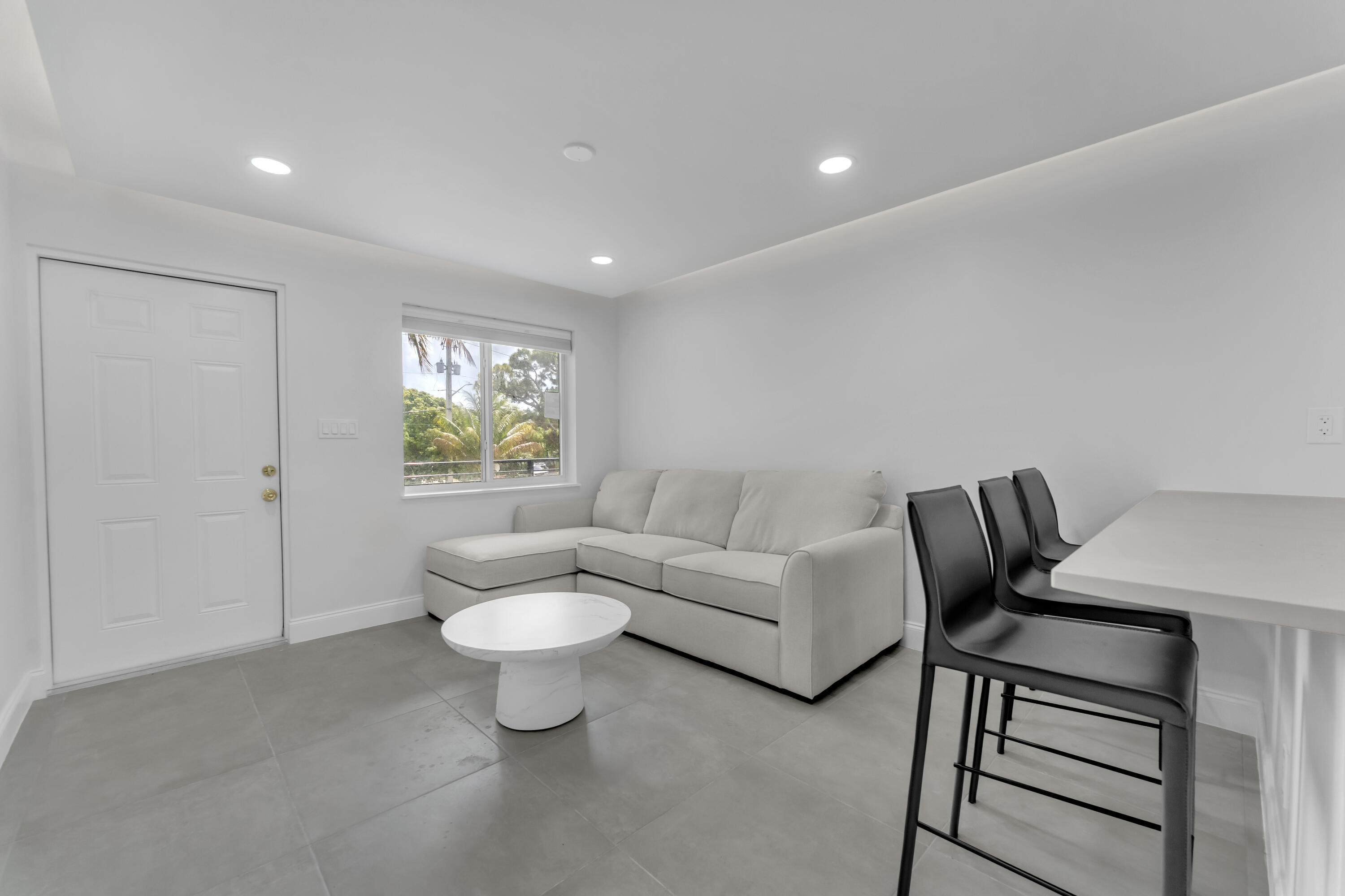 Living room with a white sectional, round coffee table, and dining table beside a palm-framed window.