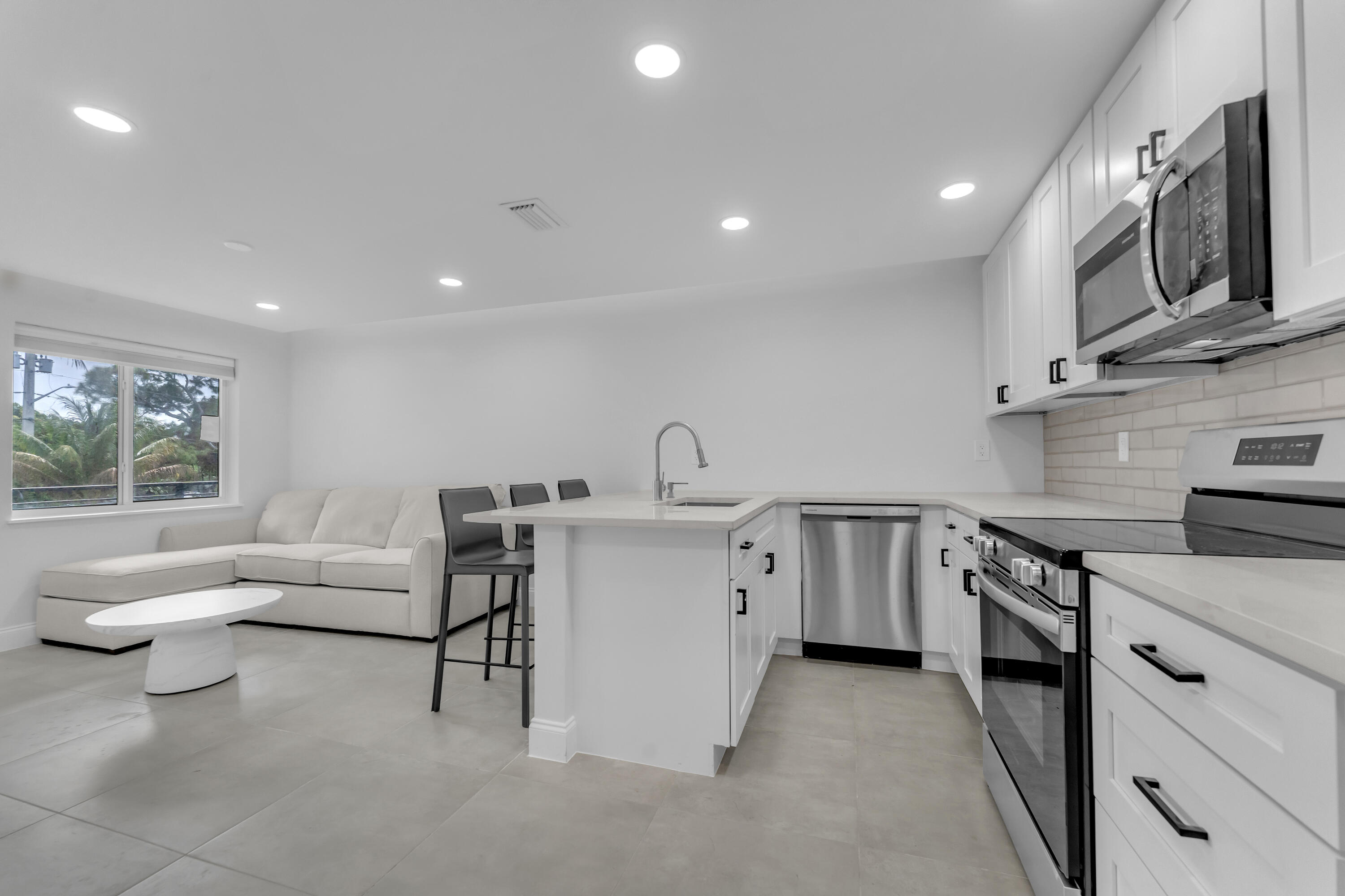 Open kitchen and living area with a white sectional, island sink, stainless appliances, and palm trees outside.