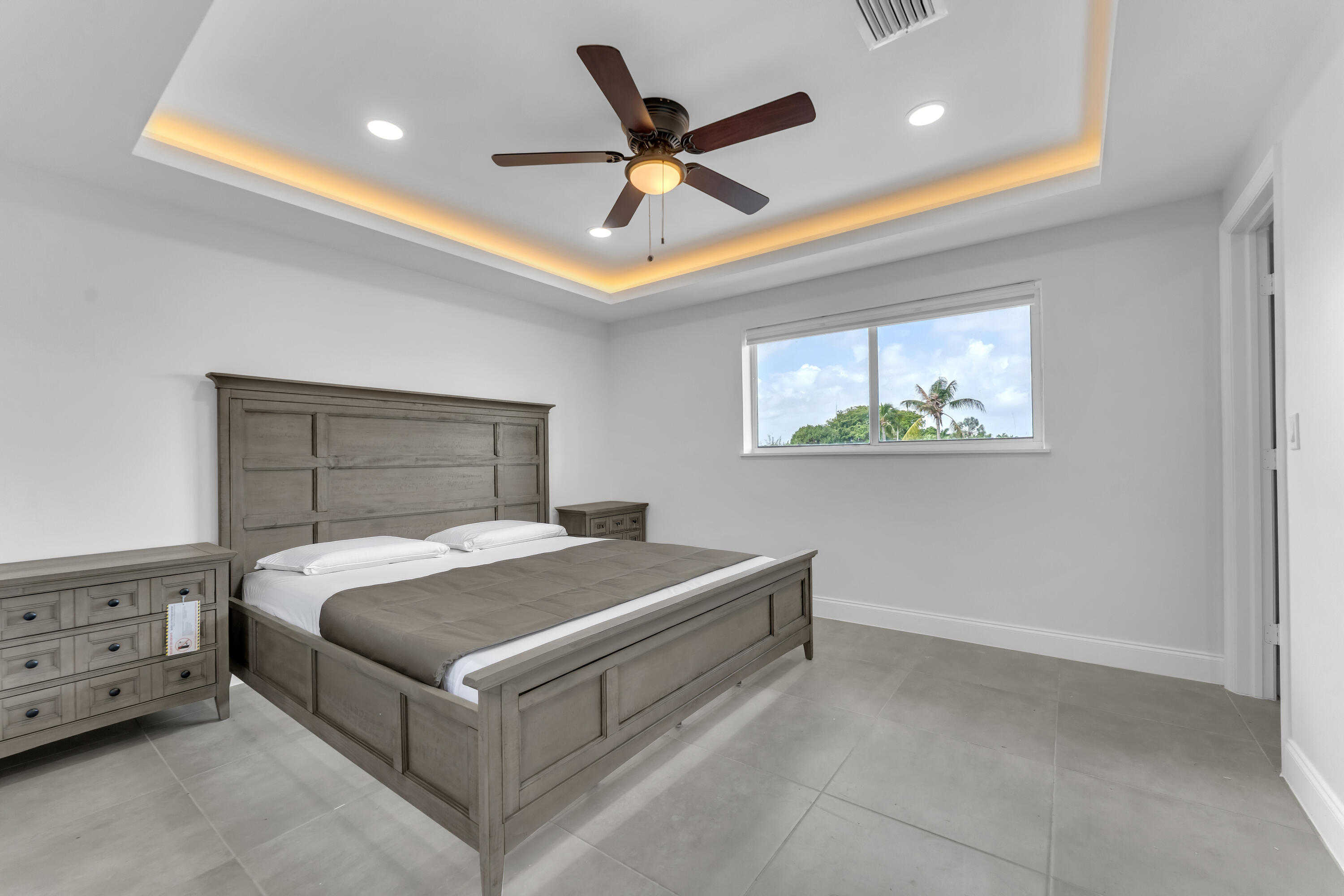 Furnished bedroom with gray wood bed, tray ceiling lighting, and palm trees visible through the window.