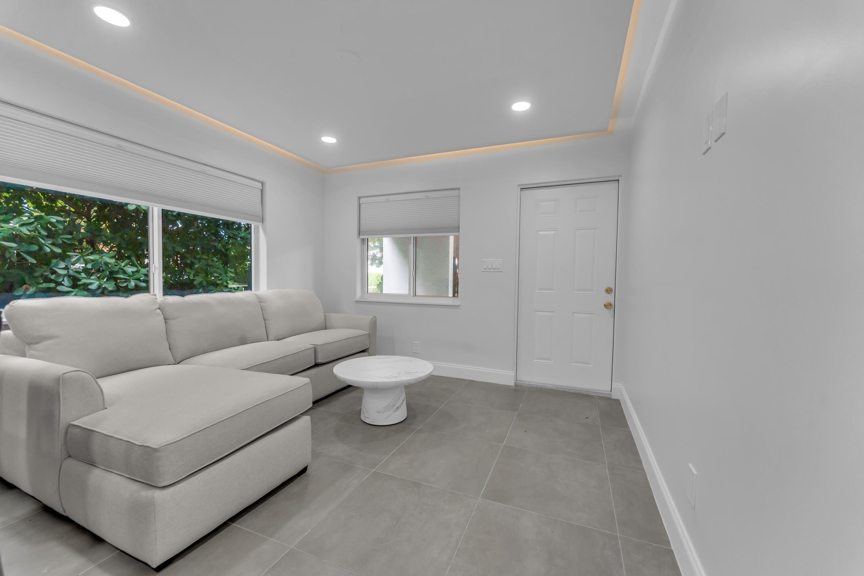 Living room with a light gray sectional, round coffee table, large windows, and gray tile floors.