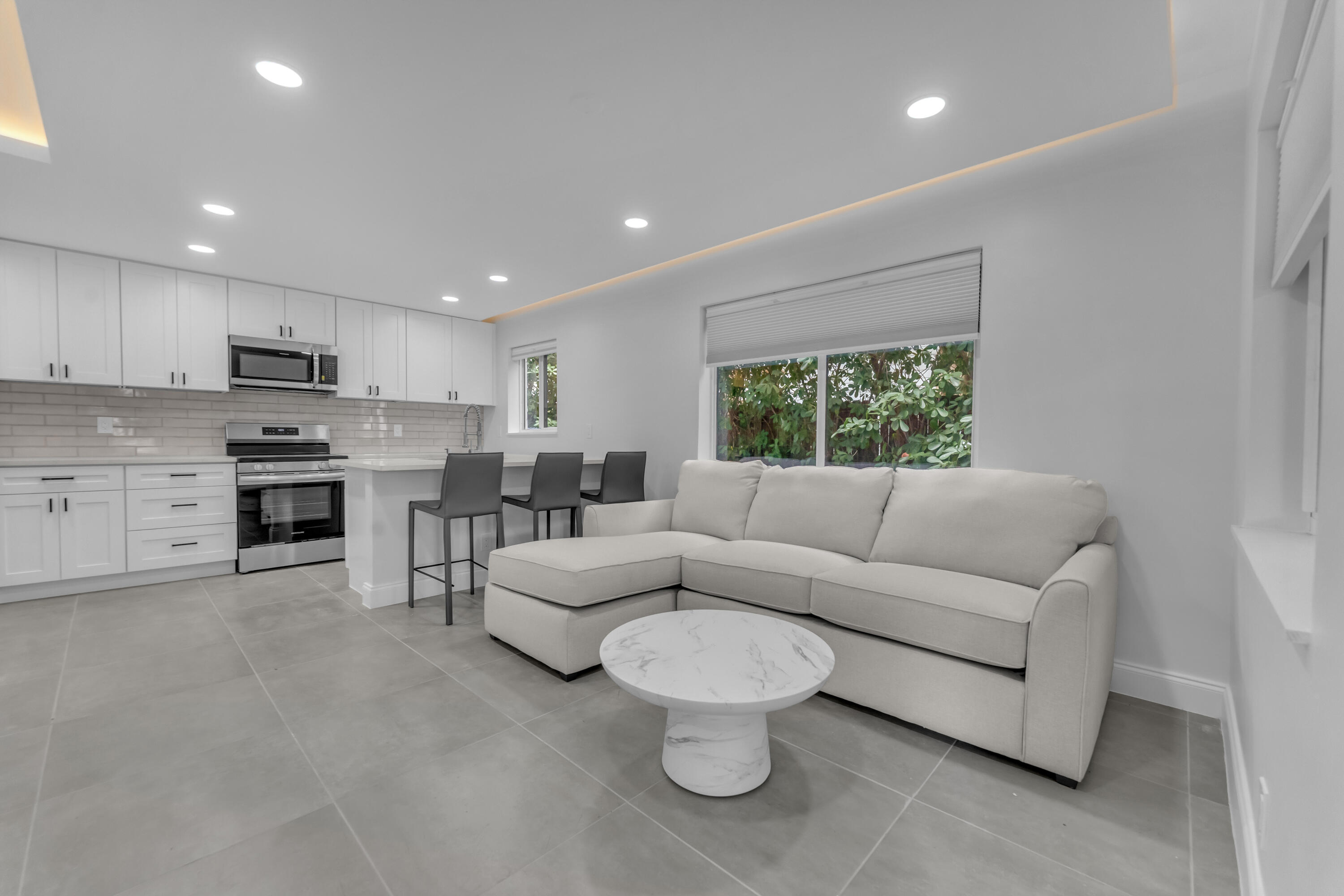 Open-plan living room and kitchen with a light sectional, breakfast bar, and greenery outside the window.