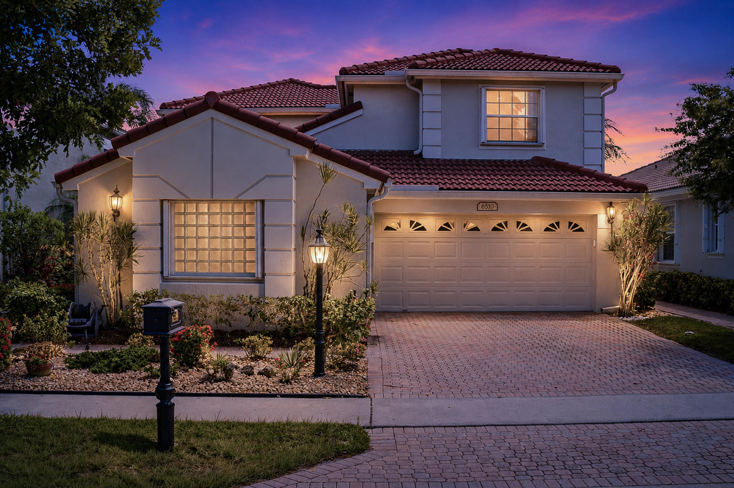 Two-story stucco house with red tile roof and illuminated windows at dusk.