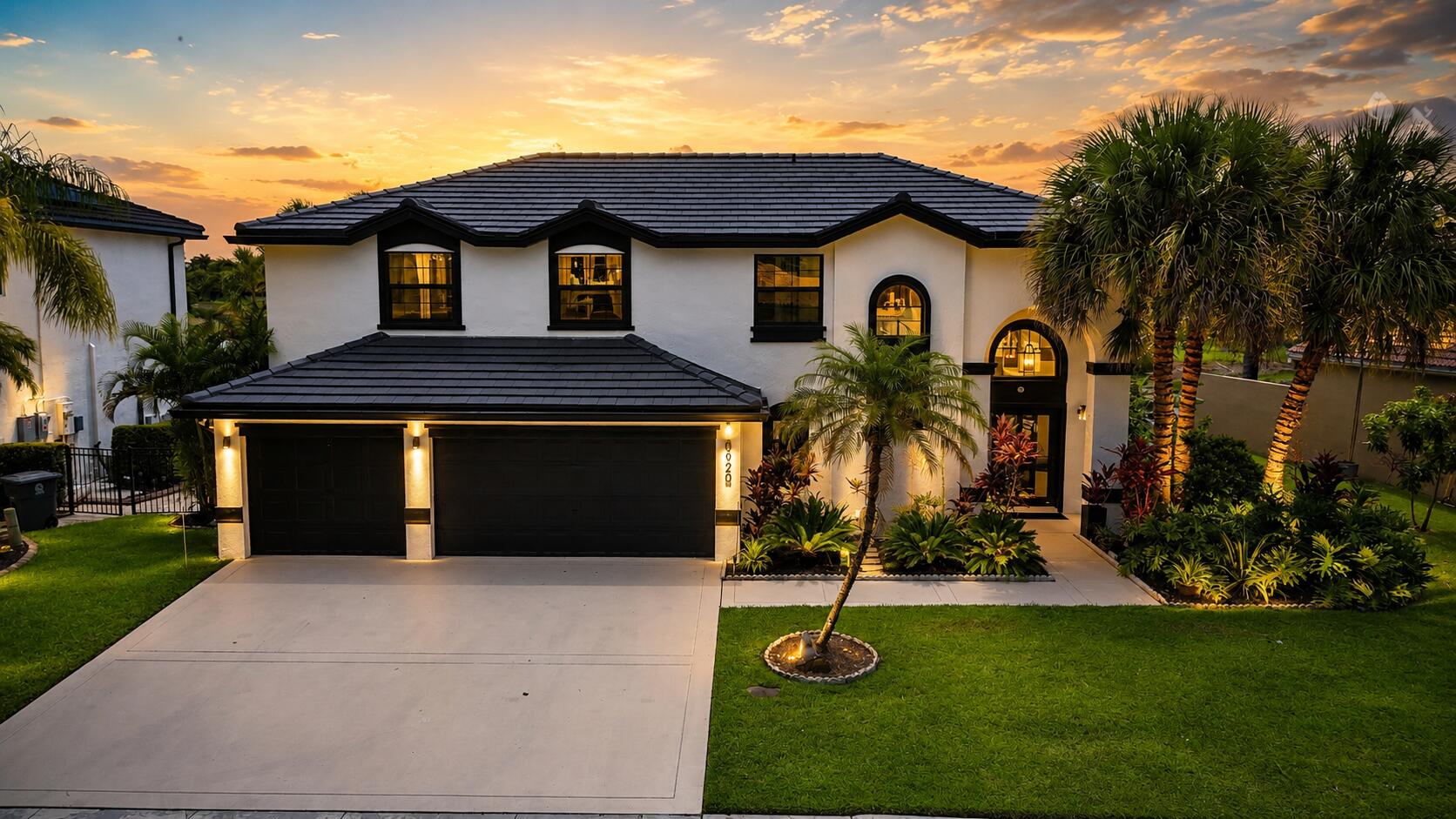 Two-story white stucco house with a dark tile roof, arched entry, and tropical landscaping at dusk.