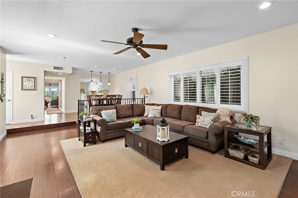 Living room with a brown sectional, plantation shutters, ceiling fan, and light wood flooring.