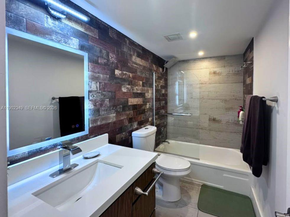 Modern bathroom with a white vanity, illuminated mirror, glass shower screen, and contrasting dark tile wall.