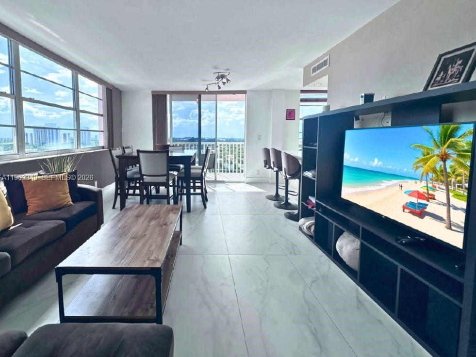 Furnished condo living and dining area with expansive windows overlooking water and a distant skyline.