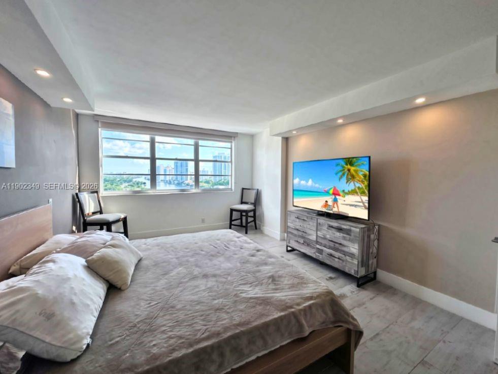Furnished bedroom with a large bed and wide window overlooking nearby buildings.