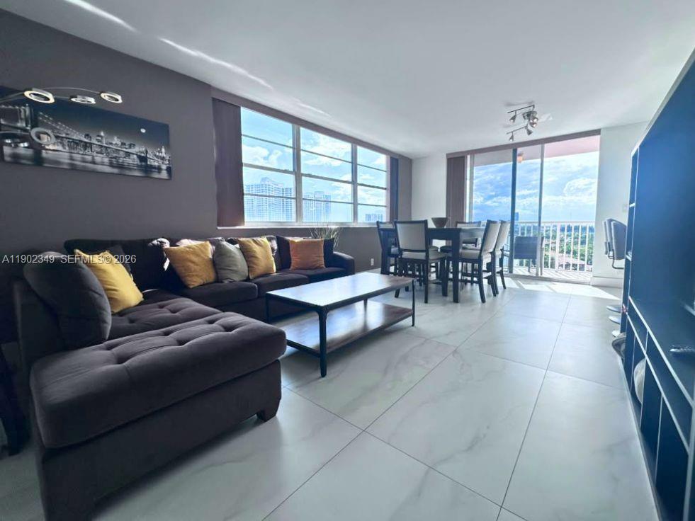Living and dining room with a dark sectional sofa, light tile floors, and large balcony windows.