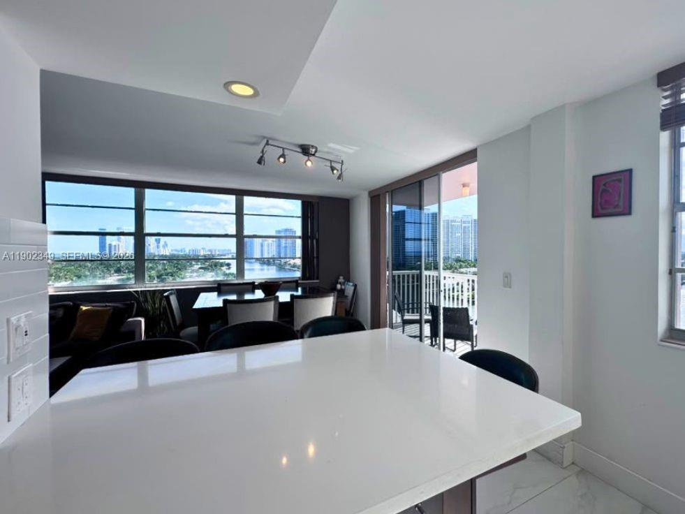 Condo kitchen and dining area with a white island, balcony, and views of water and high-rises.