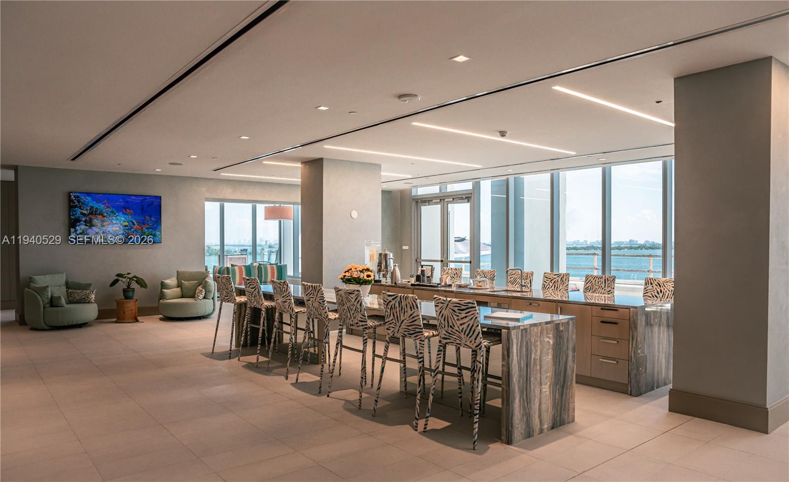Dining and lounge area with a long table and patterned chairs beside water-view windows.
