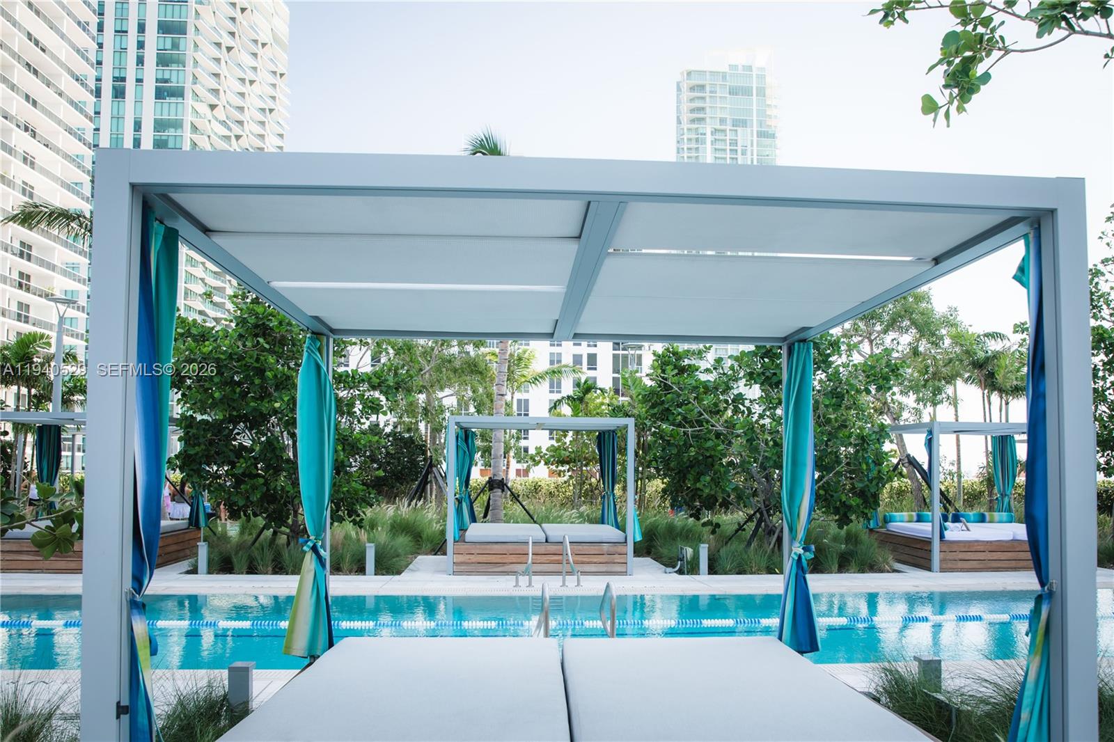Outdoor pool with curtained cabanas, white lounge cushions, and high-rise buildings in the background.