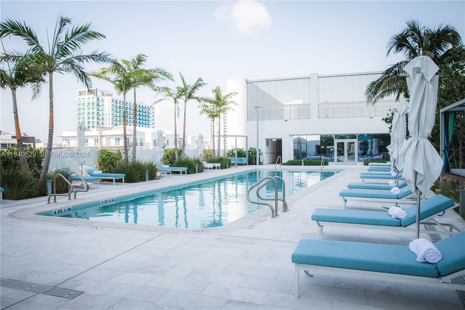 Condominium pool bordered by turquoise lounge chairs, closed umbrellas, and palm trees.