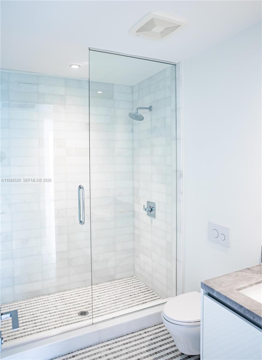 Bathroom with a glass-enclosed tiled shower, mosaic floor, wall-mounted toilet, and partial vanity.