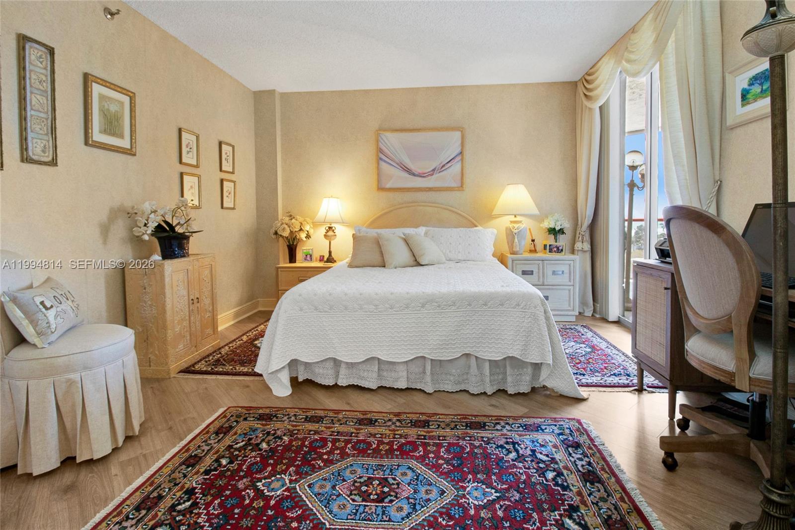 Furnished bedroom with a white bed, ornate patterned rugs, and curtained balcony doors.