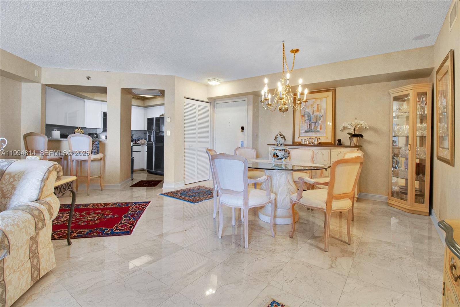 Dining area with a glass table, chandelier, marble floors, and adjoining kitchen.