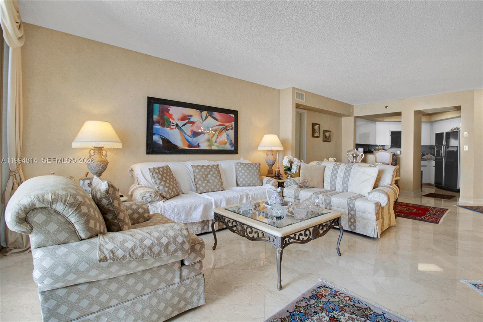 Furnished living room with cream patterned sofas, glass coffee table, and open kitchen view.