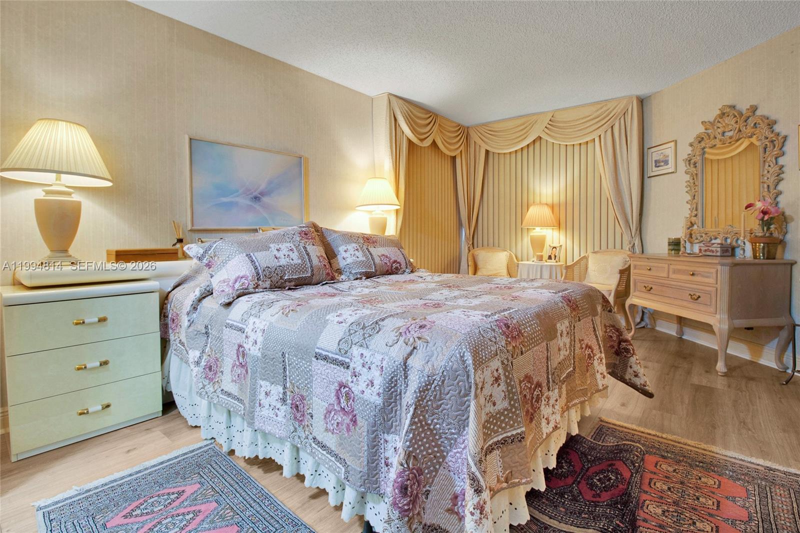 Bedroom with a floral patchwork bedspread, layered gold curtains, carved mirror, and patterned rugs.