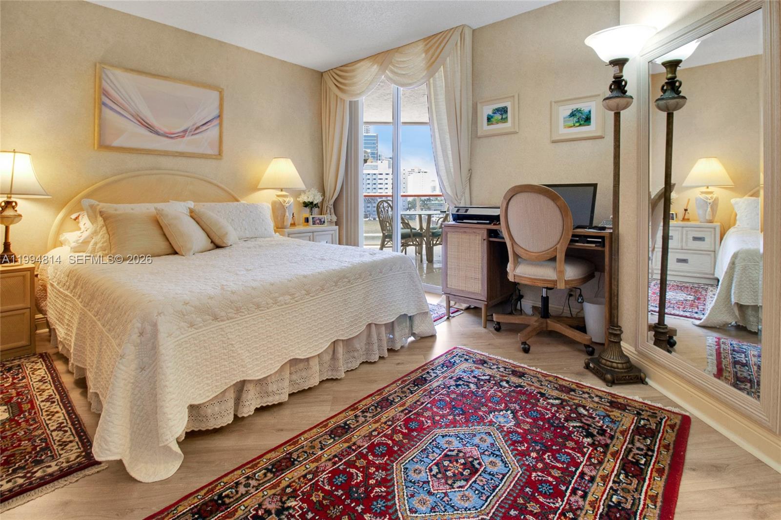 Bedroom with a white quilted bed, colorful patterned rugs, and a balcony door overlooking nearby buildings.