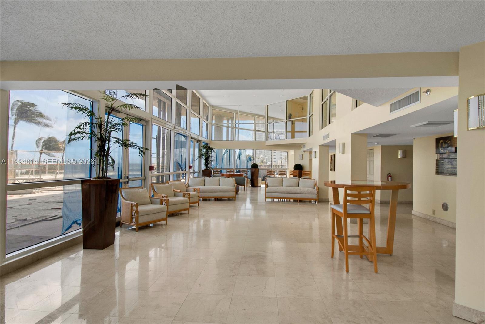 Bright condo lobby with high windows, cream sofas, potted palms, and an ocean-facing terrace.