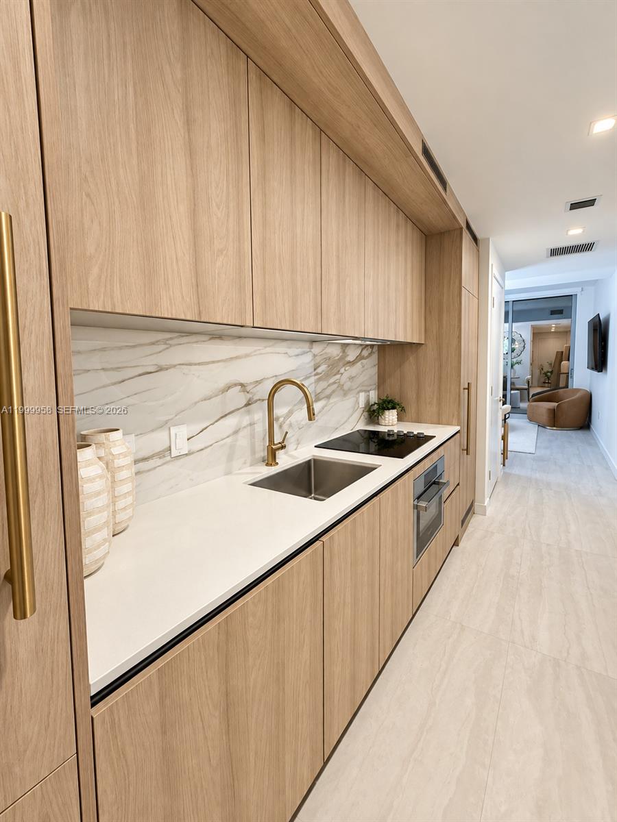 Modern condo kitchen with light wood cabinets, marble-pattern backsplash, brass faucet, and black cooktop.