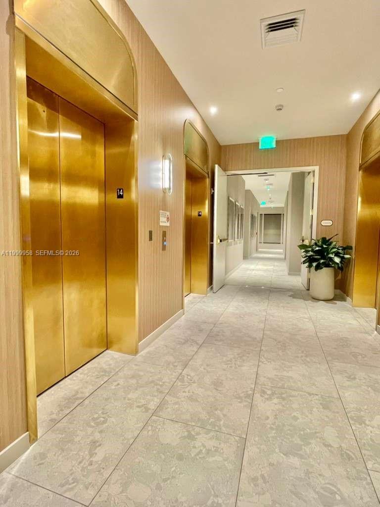Elevator lobby with gold-toned doors, pale stone tile, and a potted plant beside the corridor.