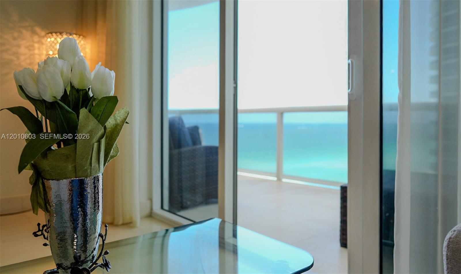 Furnished interior with white flowers beside glass doors opening onto a turquoise ocean view.