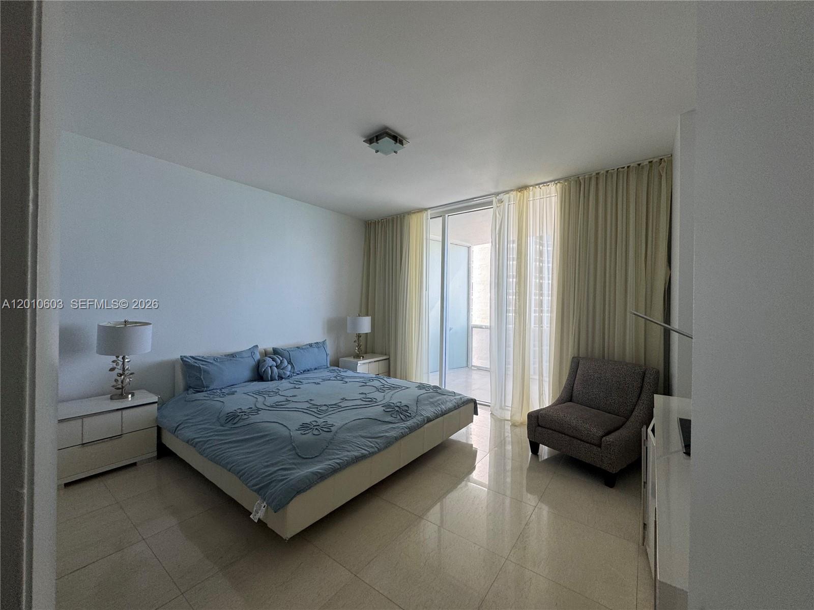 Furnished bedroom with blue bedding, a gray armchair, and sliding glass doors framed by curtains.
